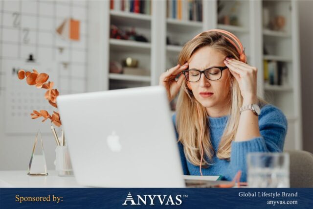 Woman sitting at a desk with her hands on her temples, looking stressed while working on a laptop, with a headset around her neck. Sponsored by Anyvas, Global Lifestyle Brand.