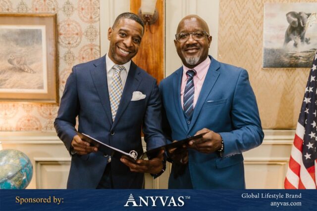 Two professionally dressed men smiling and holding documents, standing in a formal setting with a globe, artwork, and an American flag in the background. Sponsored by Anyvas, a global lifestyle brand.