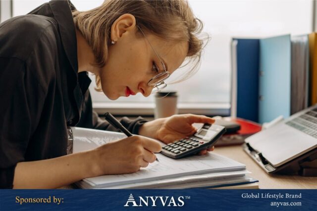 A focused woman wearing glasses works diligently at her desk, holding a calculator in one hand and writing in a notebook with the other. She is surrounded by a laptop, office supplies, and organized folders, representing a professional engaged in detailed auditing or financial tasks.