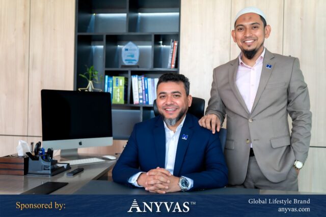 Hasan Imam Siddiki seated confidently in his office chair, with Shah Md. Mohin Uddin standing beside him, both smiling warmly. The office backdrop features bookshelves and professional decor. Sponsored by ANYVAS.