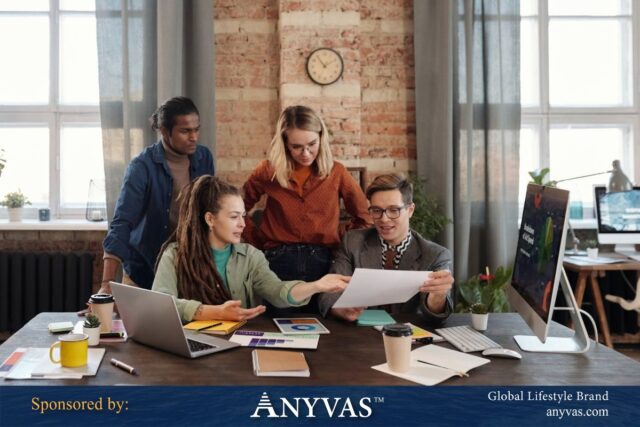 A diverse group of four professionals collaborating in a modern office, reviewing documents and digital reports on a desk filled with laptops, papers, and coffee cups. The scene is sponsored by ANYVAS, a global lifestyle brand.